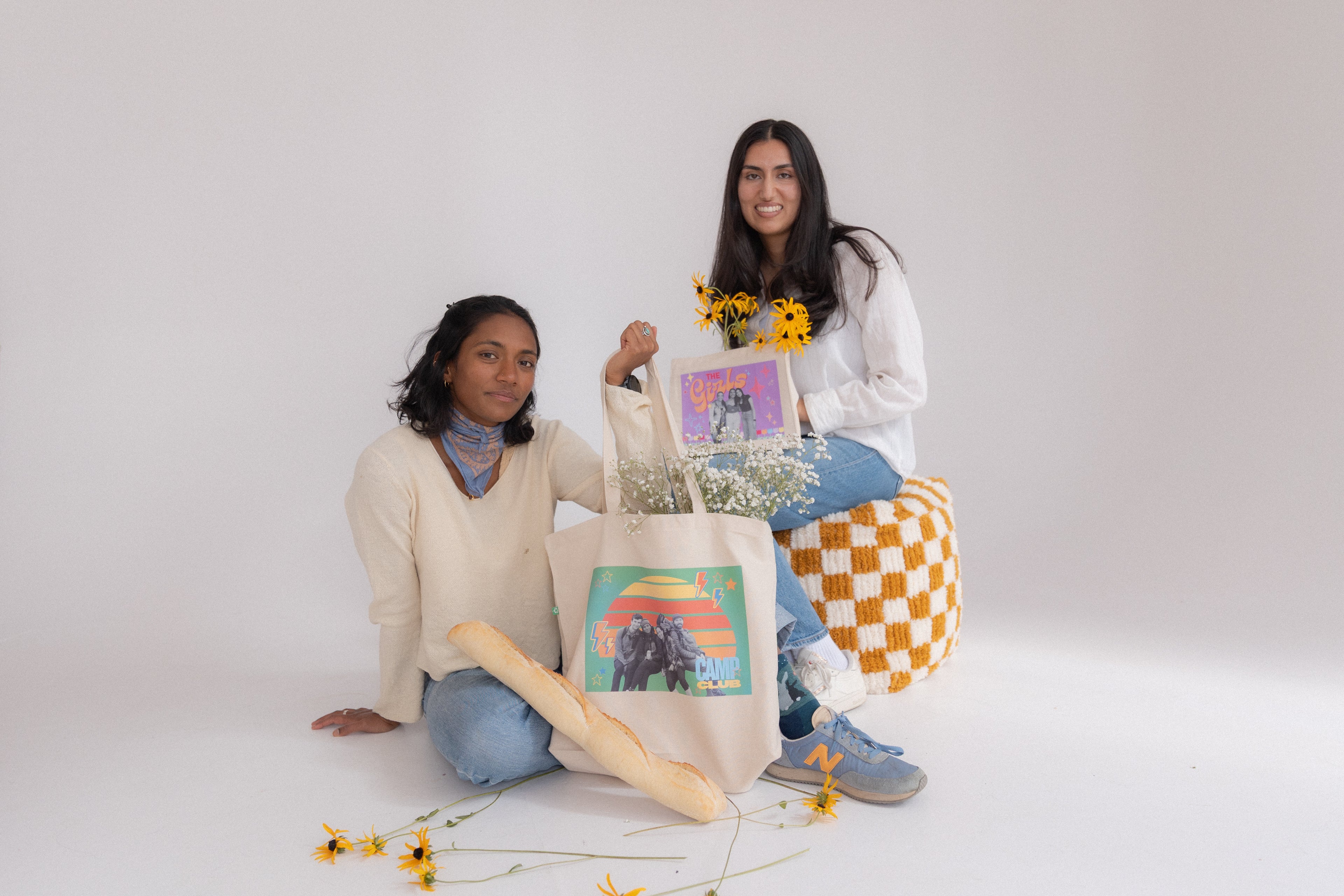 Two women sitting on a white floor with colorful tote bags and flowers.