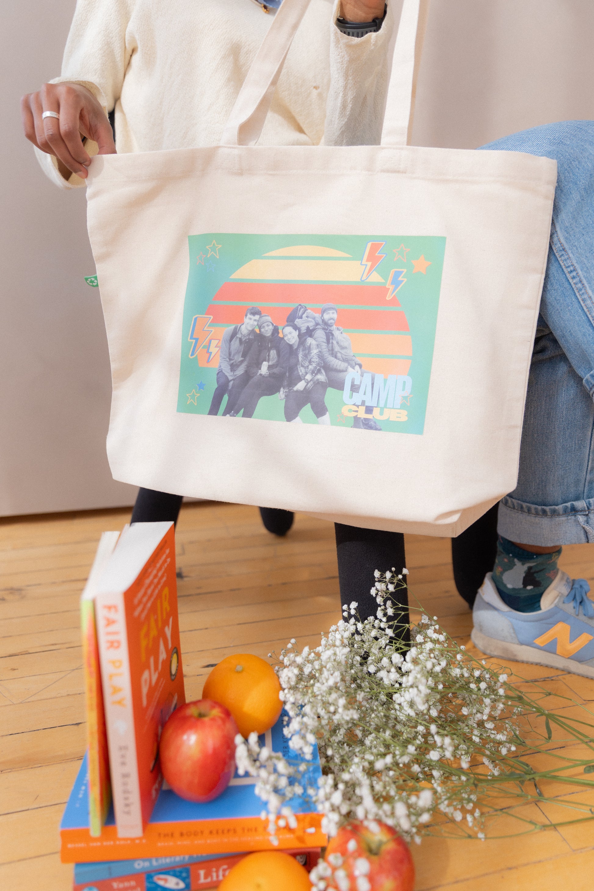 Person holding a tote bag with a colorful design, standing next to books and fruits on a wooden floor.