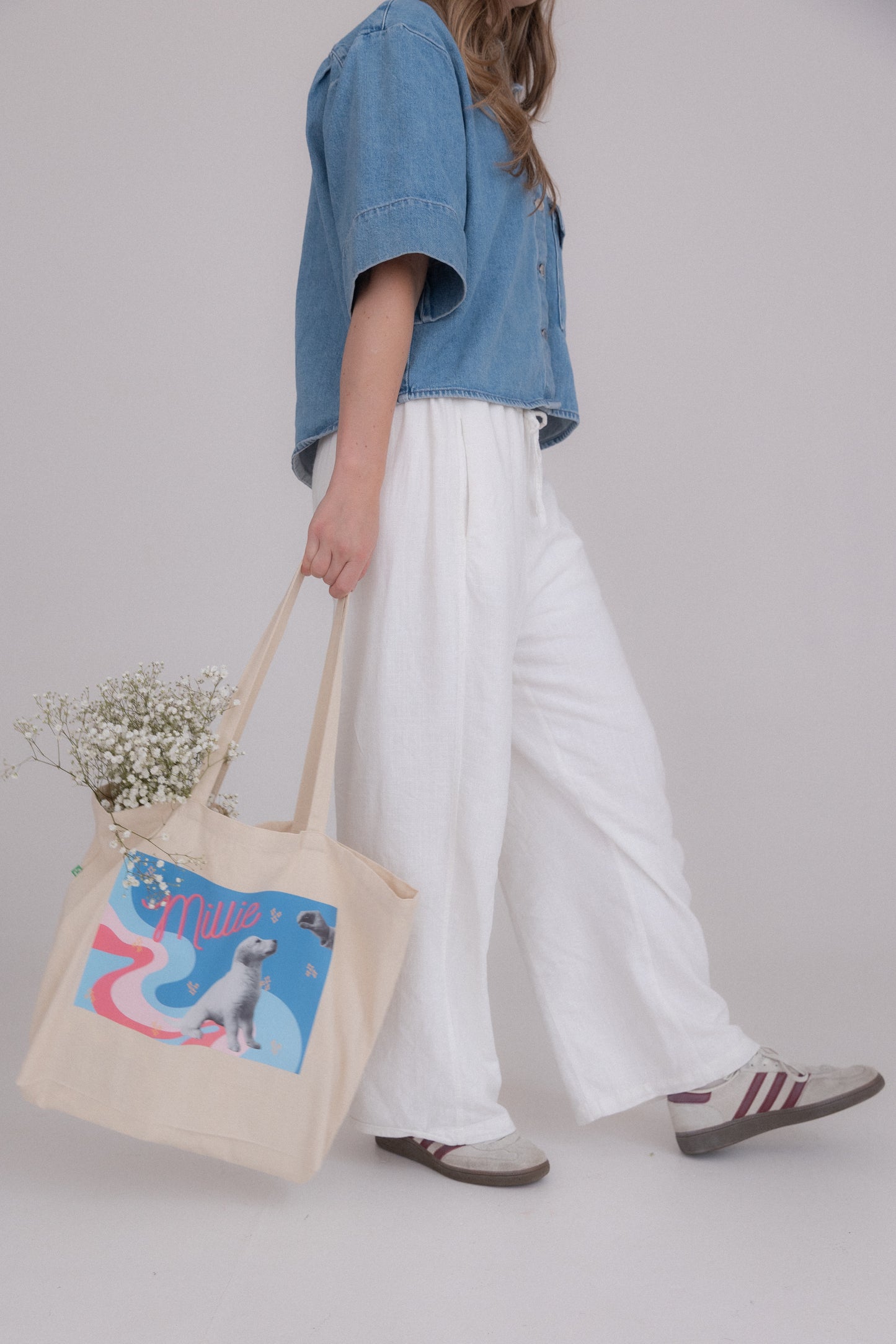 Person holding a tote bag with a design, wearing a denim jacket and white pants on a plain background