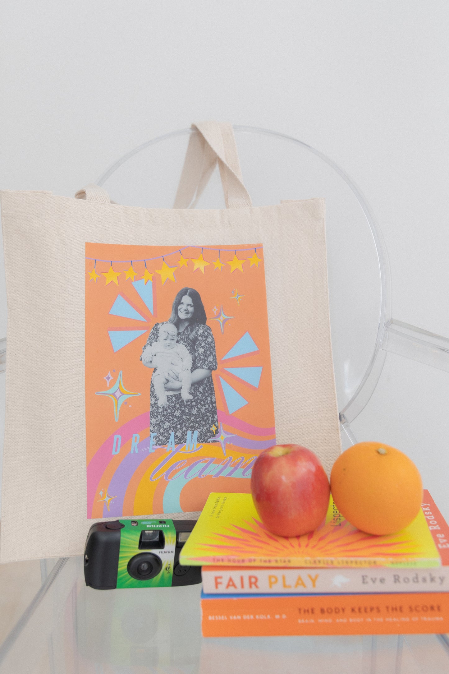 Tote bag with colorful design featuring a woman and child, placed on books with apples and an orange.