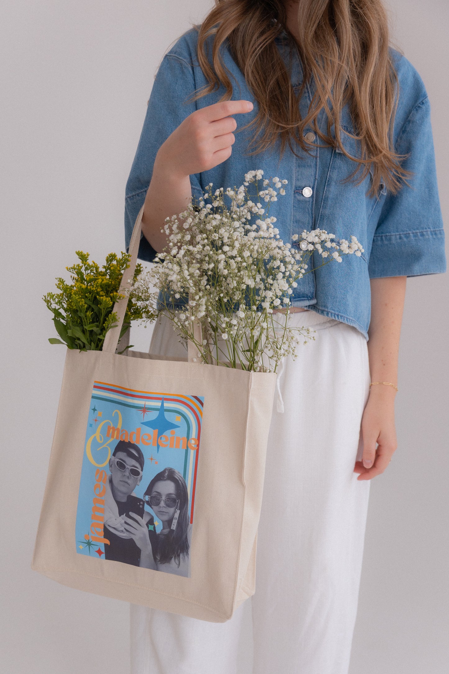 Person holding a tote bag with a graphic design and flowers, wearing a denim jacket and white pants.