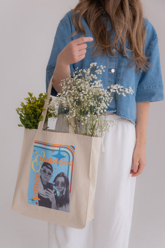 Person holding a tote bag with a graphic design and flowers, wearing a denim jacket and white pants.